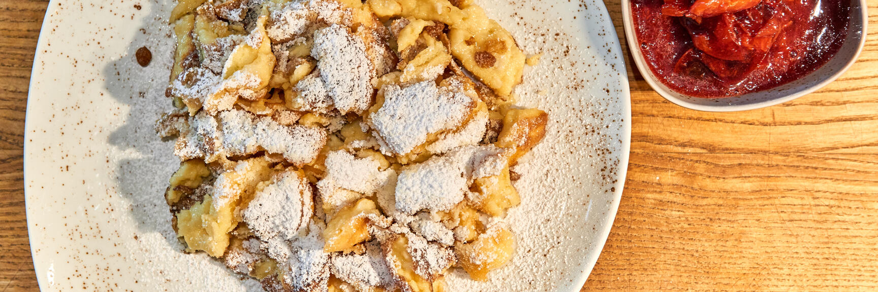 Kaiserschmarrn with powdered sugar on white plate, bowl of plum compote beside it, wooden table.
