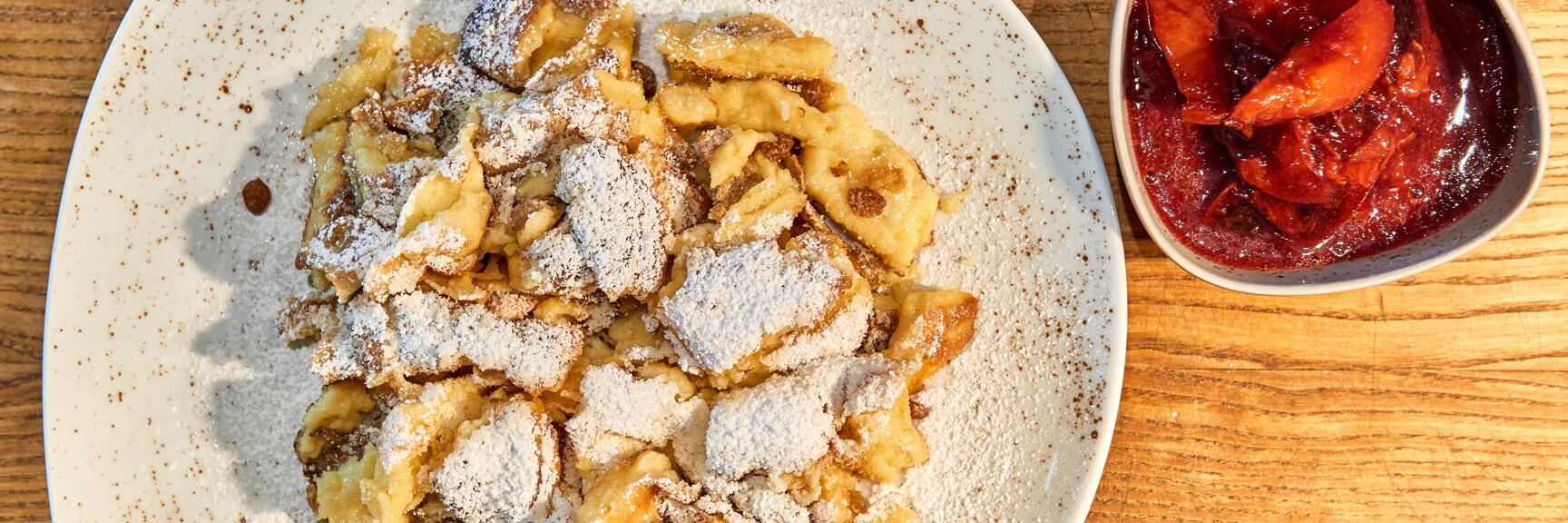 Kaiserschmarrn with powdered sugar on white plate, bowl of plum compote beside it, wooden table.