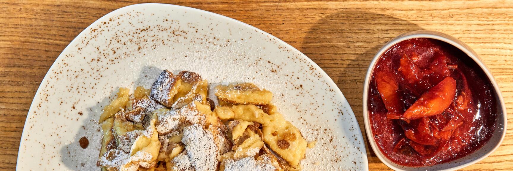Kaiserschmarrn with powdered sugar on white plate, bowl of plum compote beside it, wooden table.
