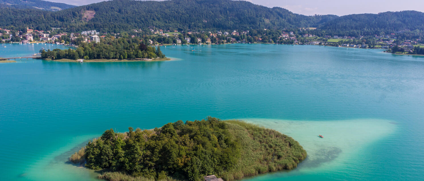 Aerial view of a small forested island in turquoise Lake Wörthersee with village and hills in background.