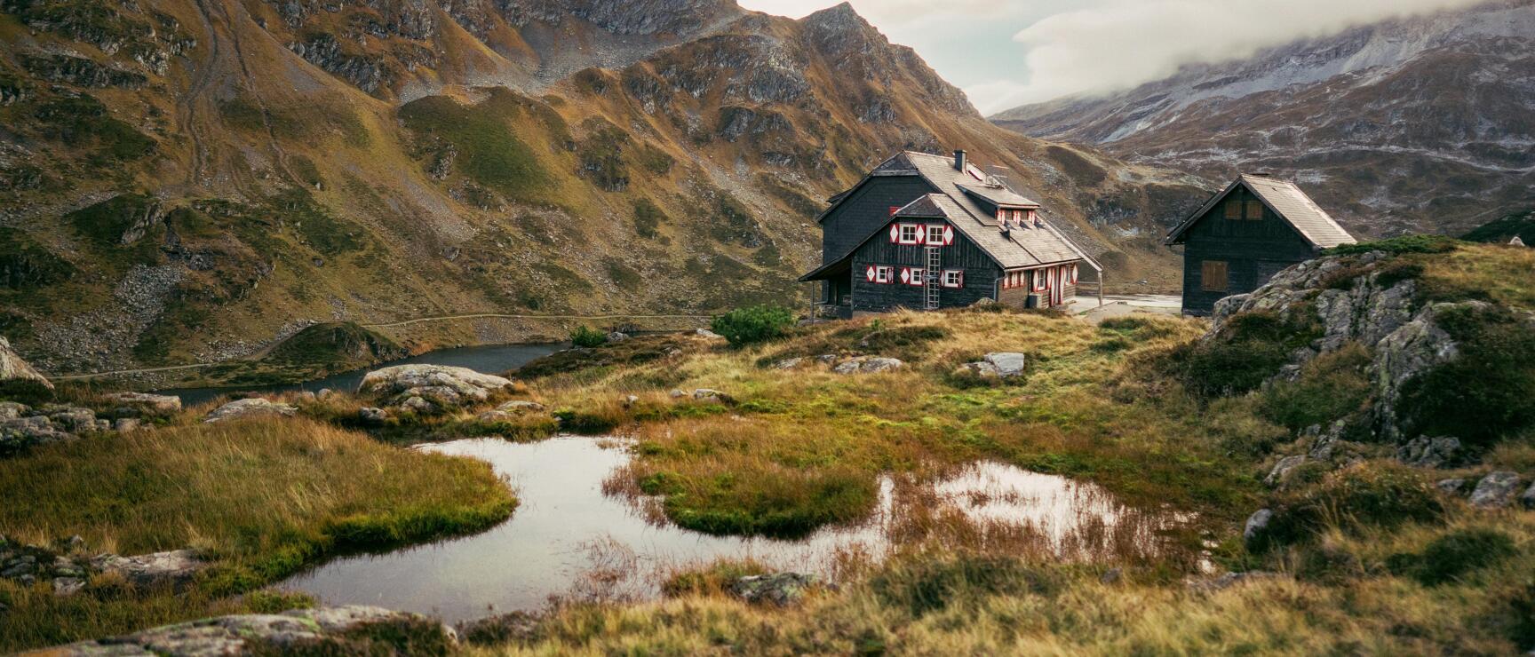 Dark alpine hut with red decorations beside a small mountain lake, rocky peaks in the background.