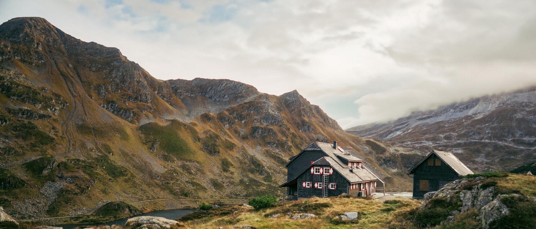 Dark alpine hut with red decorations beside a small mountain lake, rocky peaks in the background.
