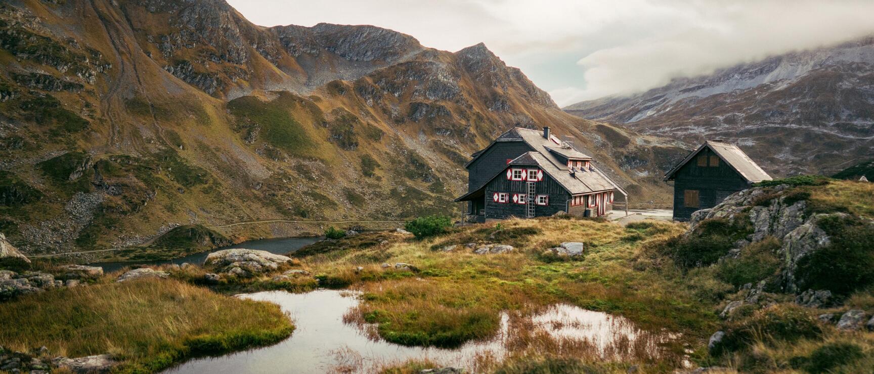 Dunkle Almhütte mit roten Verzierungen neben kleinem Bergsee, felsige Gipfel im Hintergrund.