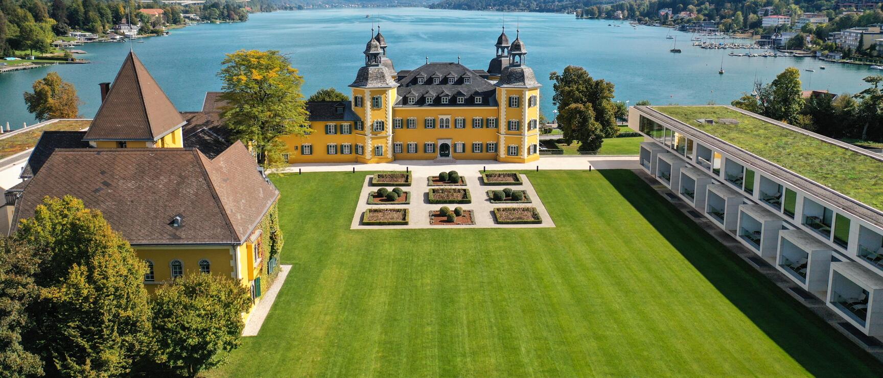Aerial view of Velden Castle on Lake Wörthersee with yellow Baroque building, gardens and lake in background.