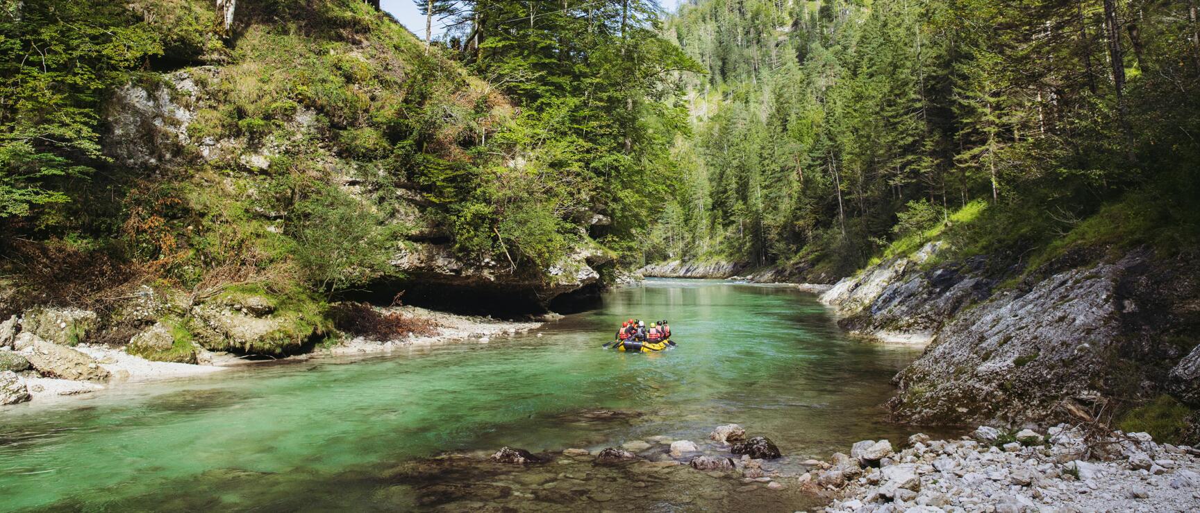 Rafting boat on turquoise river in forested canyon with suspension bridge overhead and rocky banks.