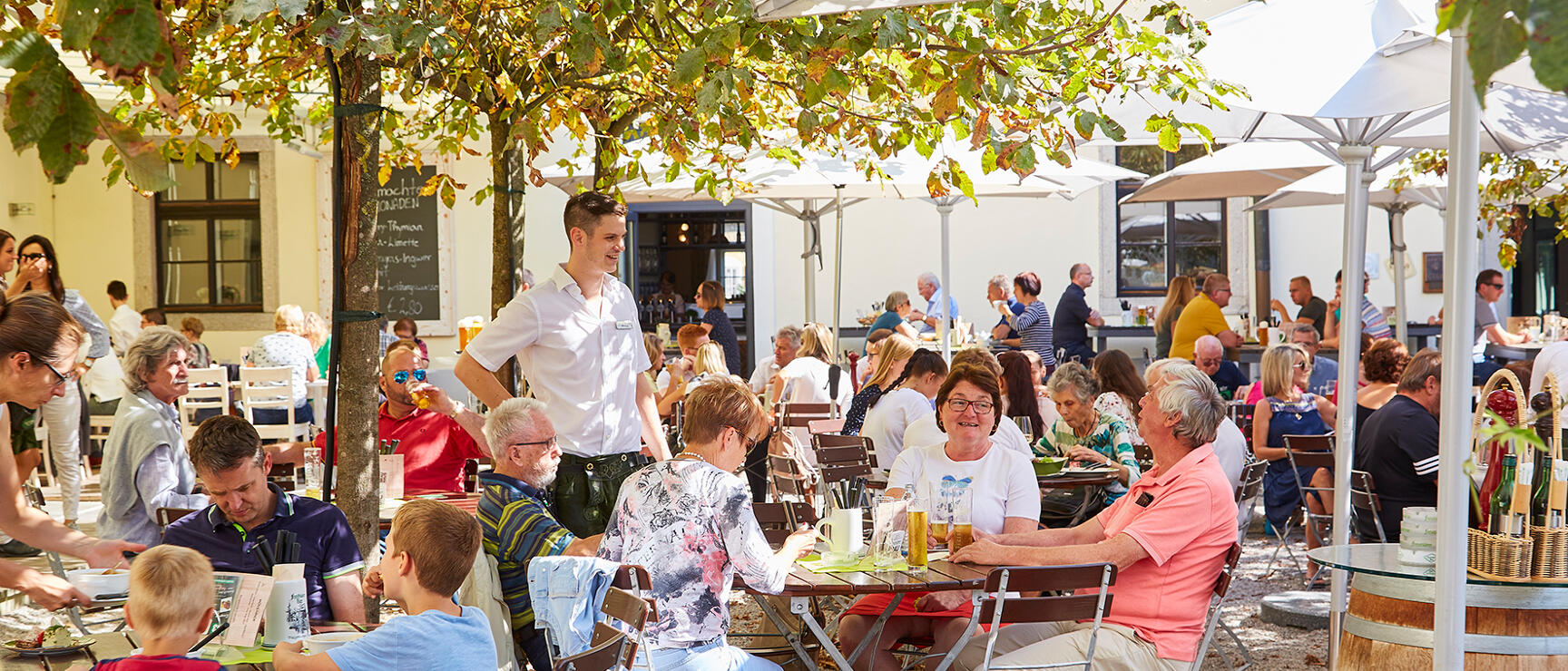 Busy beer garden with many guests at wooden tables, sun umbrellas and trees, waiter in white shirt.