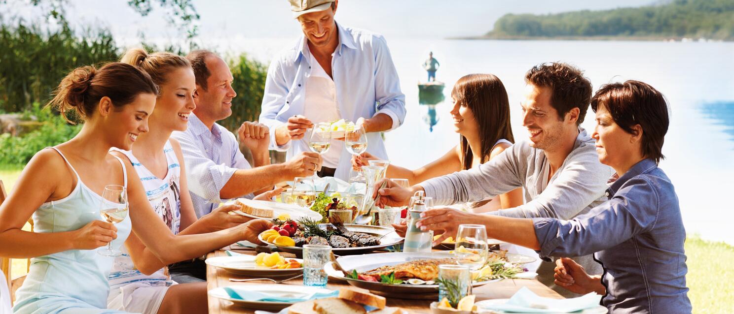 Group of seven people dining at a wooden table outdoors, wine glasses, lake and mountains in the background.