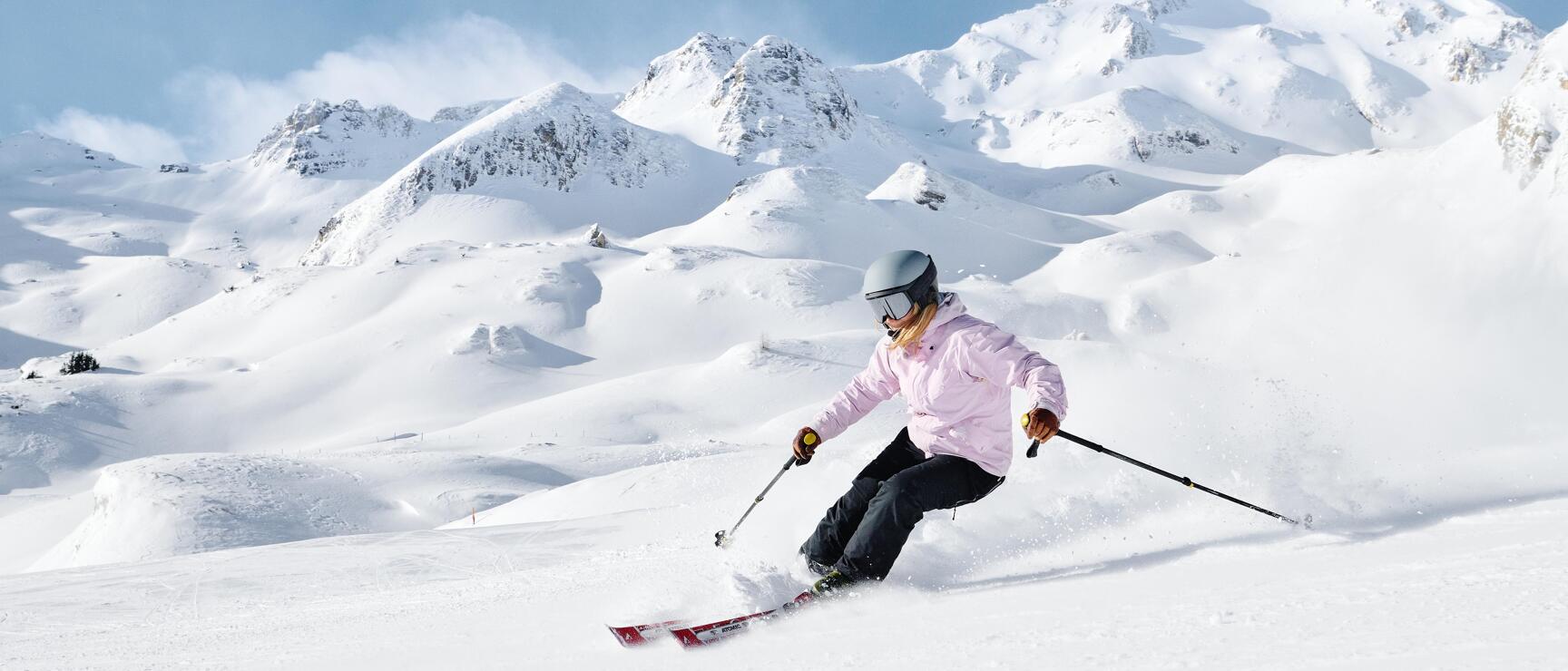 Female skier in pink jacket and grey helmet on snowy slopes, snow-capped mountain peaks in background.