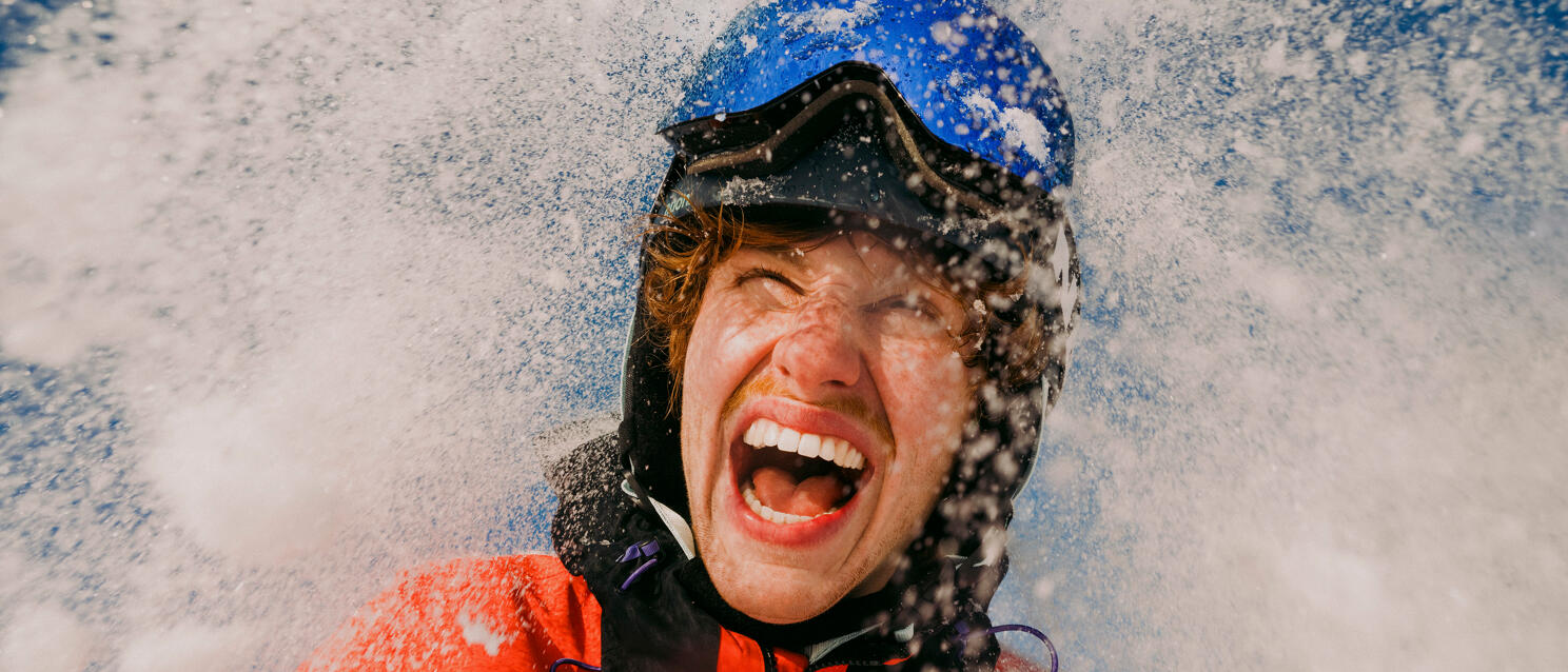 Laughing person wearing blue helmet and red jacket surrounded by splashing snowflakes.