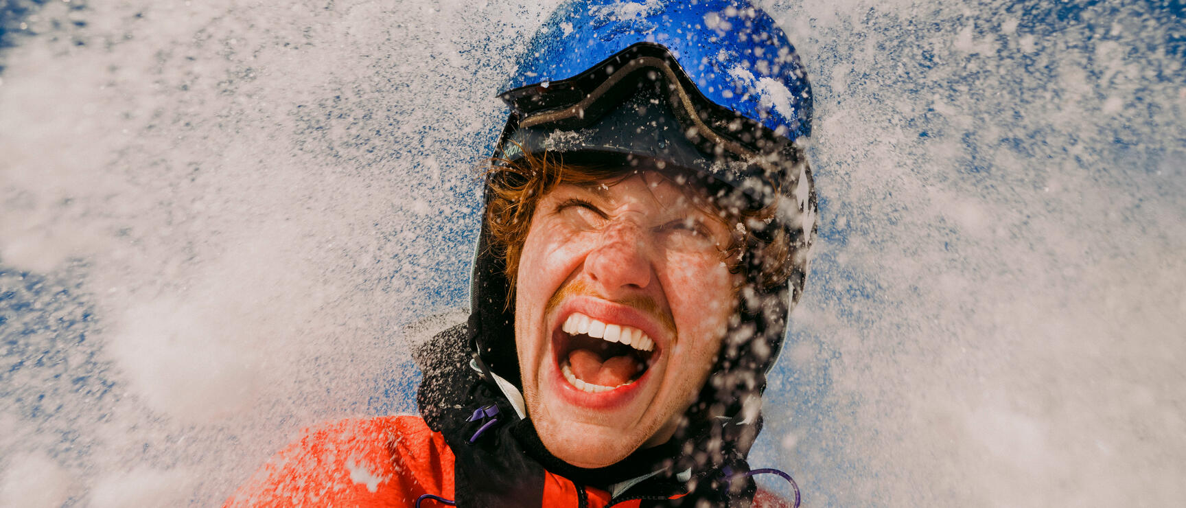 Laughing person wearing blue helmet and red jacket surrounded by splashing snowflakes.