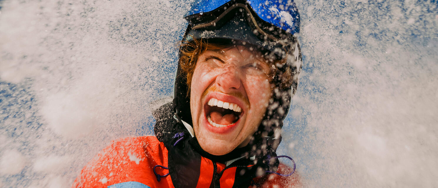 Laughing person wearing blue helmet and red jacket surrounded by splashing snowflakes.