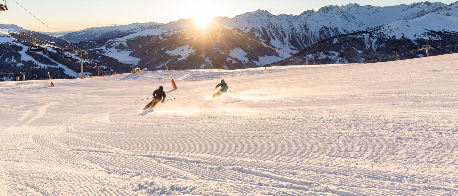 Two skiers on a groomed slope in backlighting, with a chairlift and snow-covered mountains in the background.