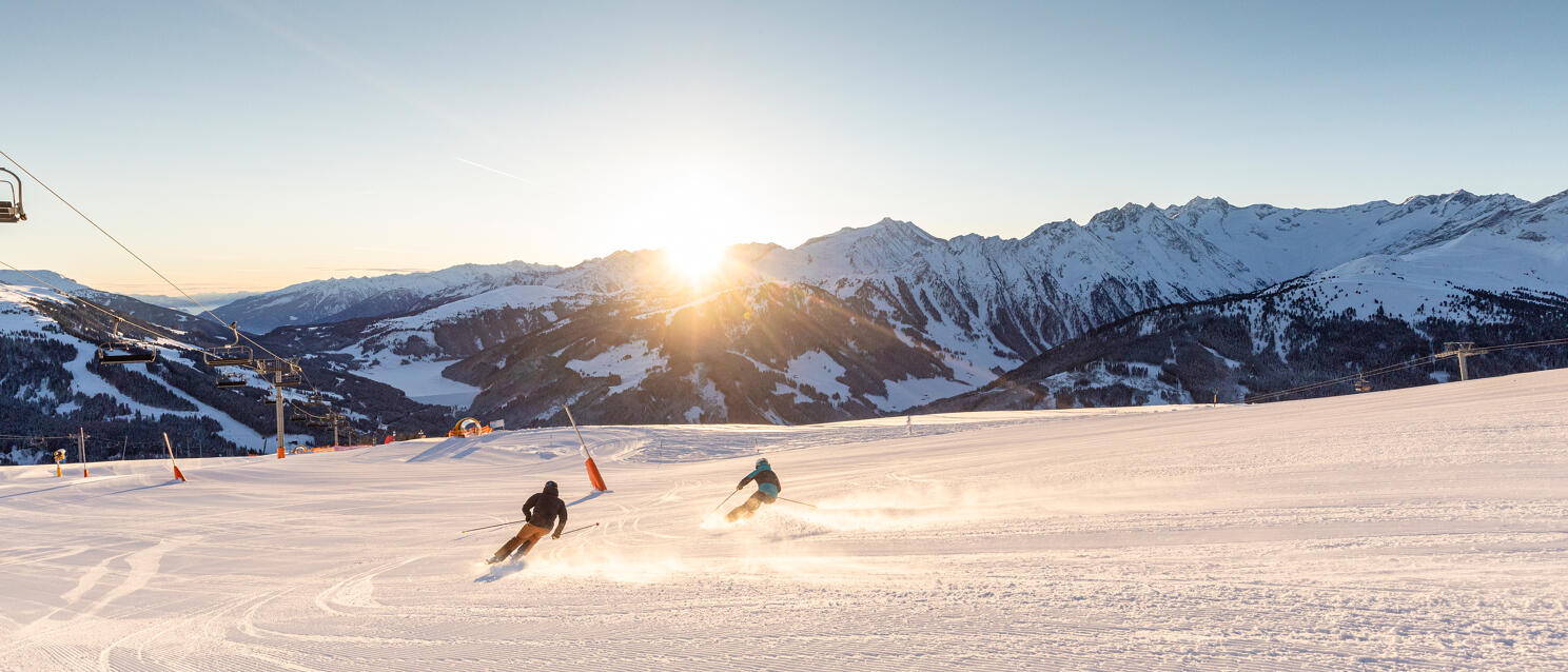 Two skiers on a groomed slope in backlighting, with a chairlift and snow-covered mountains in the background.