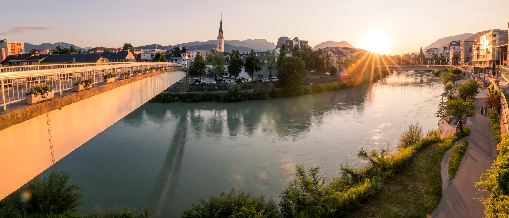 White pedestrian bridge over river at sunset with church tower and mountains in background, Villach