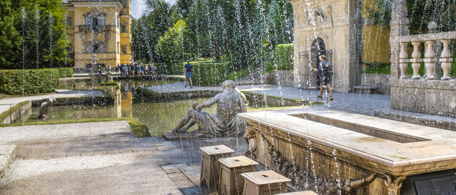 Water features with stone statue at Hellbrunn Palace, yellow castle building and visitors in the background.