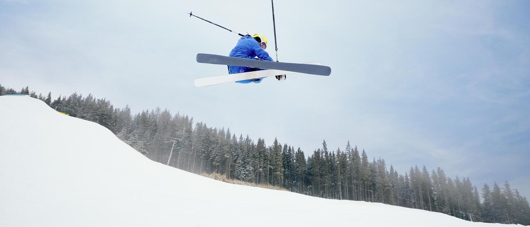 Skifahrer in blauer Jacke und gelbem Helm bei einem hohen Sprung, Nadelwald und Schnee im Hintergrund.
