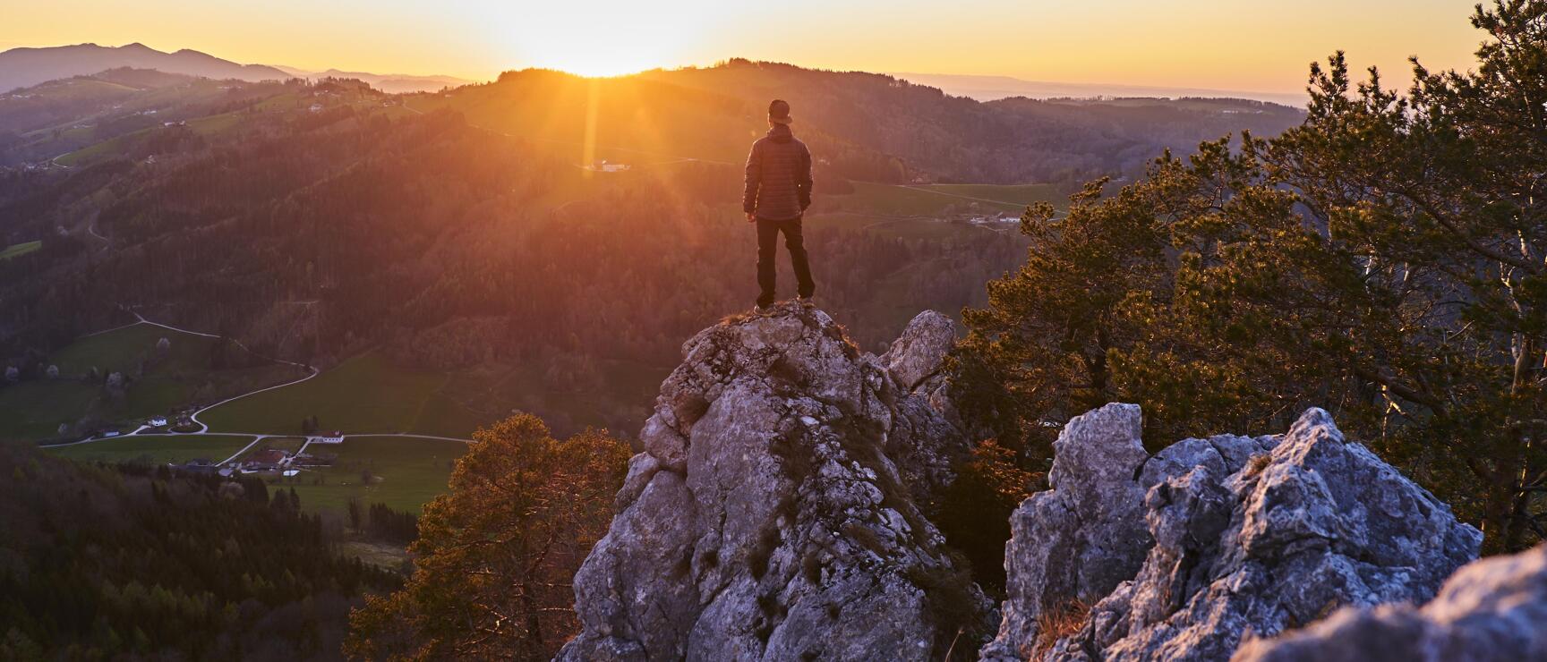Person steht auf einem Felsen bei Sonnenuntergang, Panoramablick auf Hügel und Täler im Hintergrund.