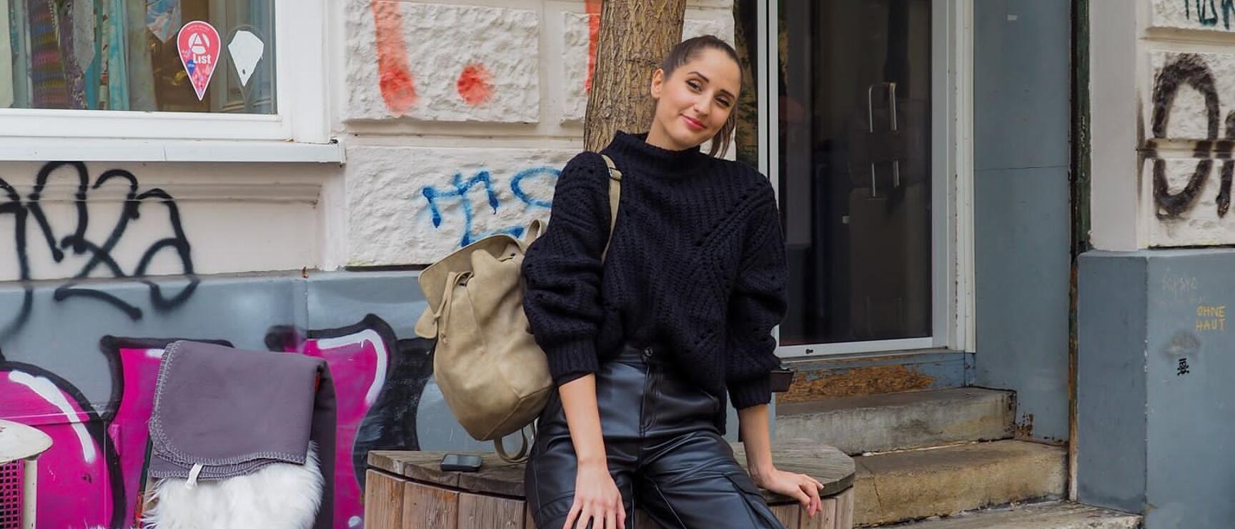 Woman in black sweater and leather pants sits on wooden post in front of graffitied building.