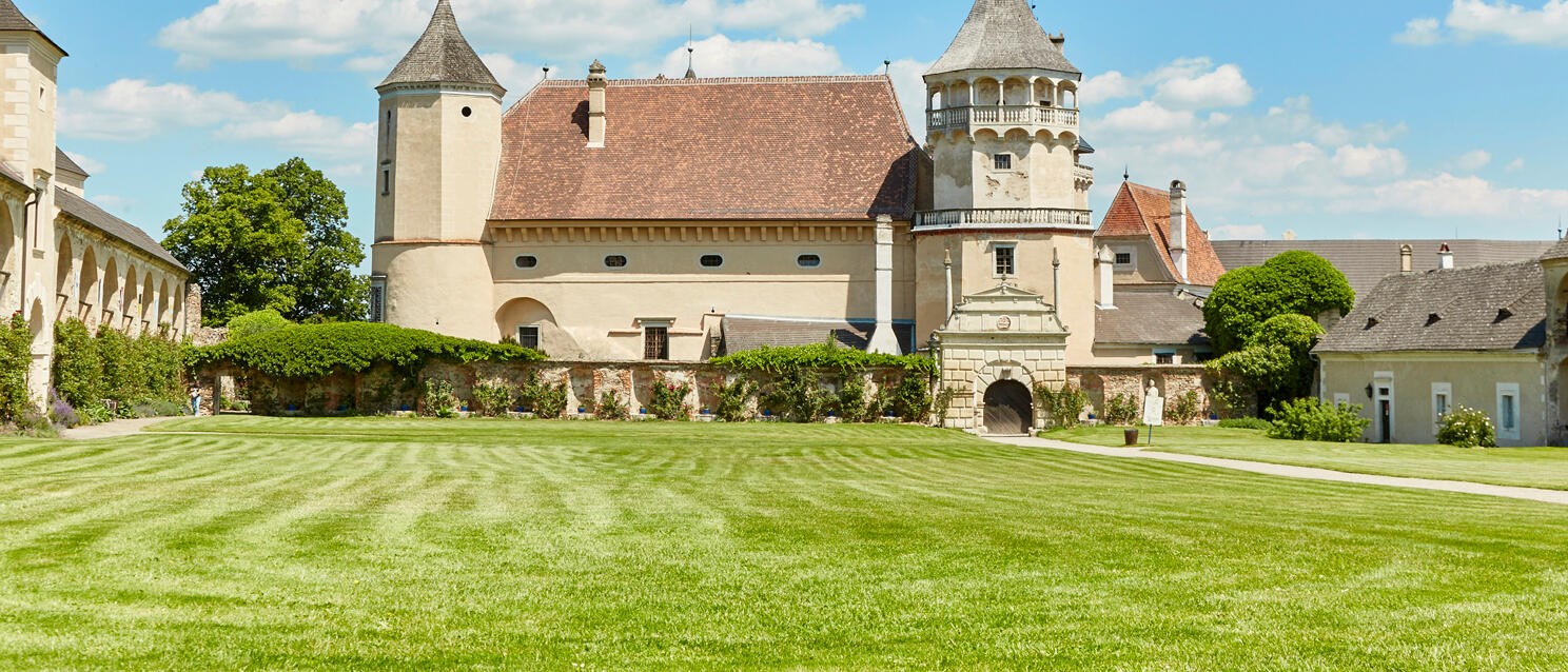 Castle with two towers and red tiled roof, manicured lawn in foreground, blue sky.