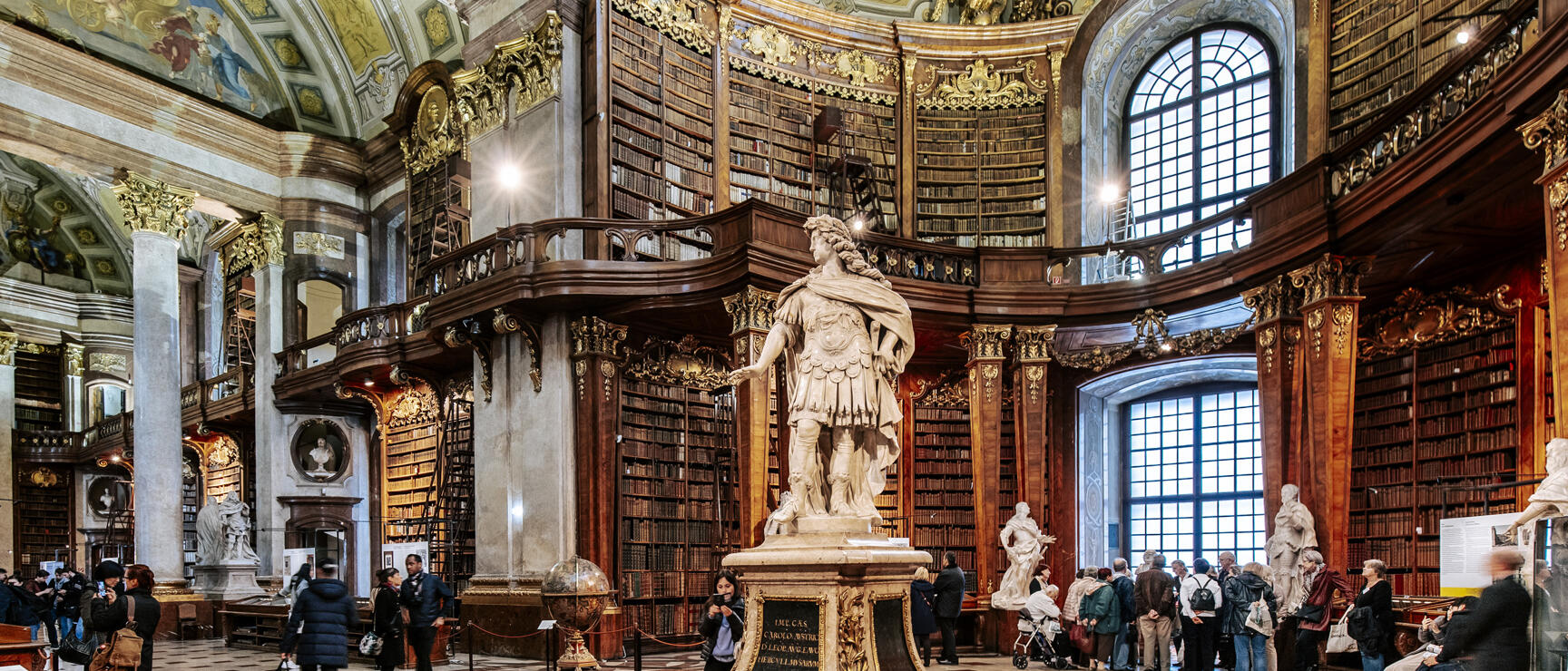Grand Hall of the Austrian National Library Vienna, marble statue in centre, visitors, bookshelves around.