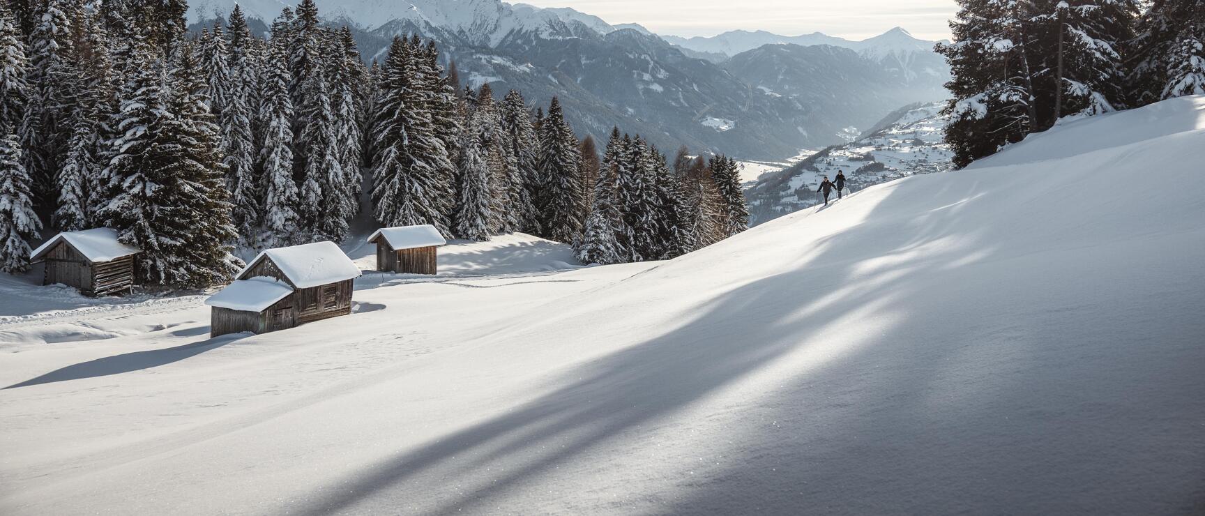 Snow-covered alpine landscape with wooden huts, two people on snowshoes, mountain panorama in the background.