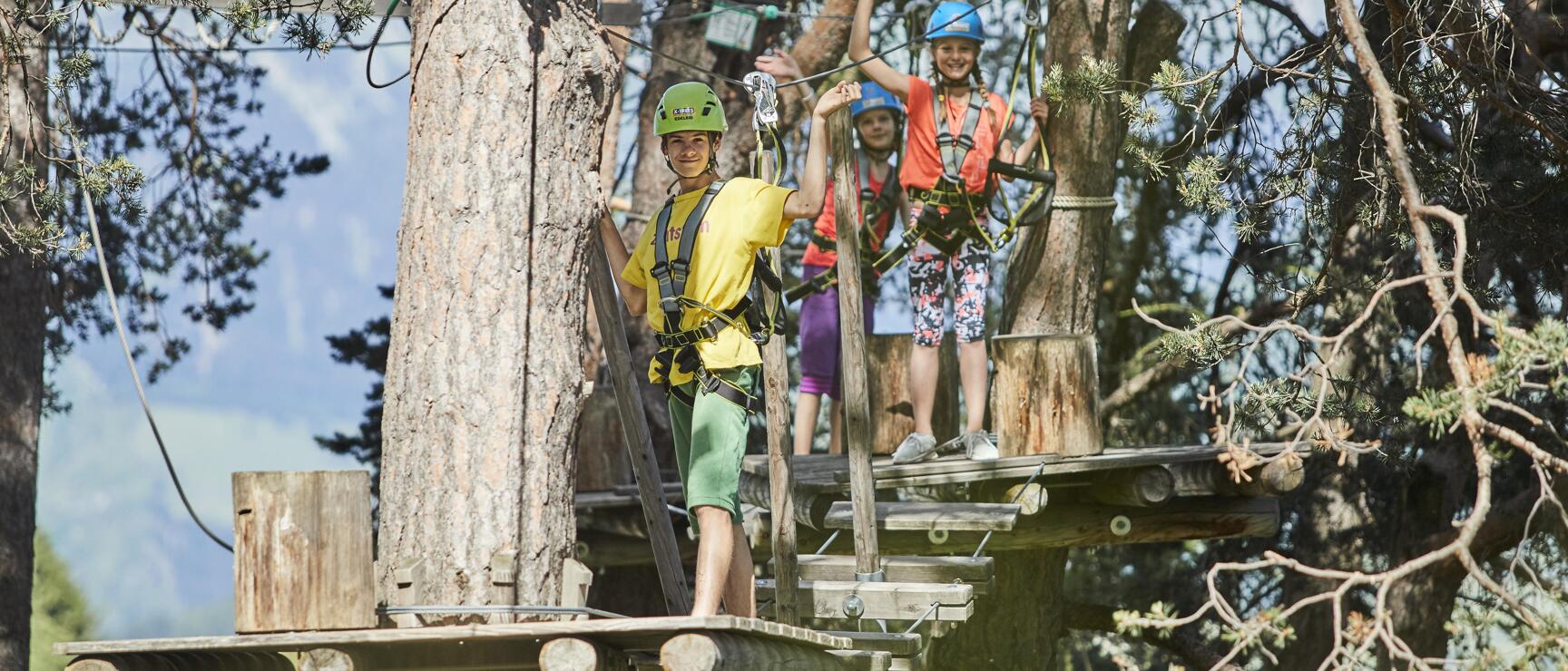 Three children with helmets and climbing harnesses on wooden platforms in a high rope course, forest in the background.