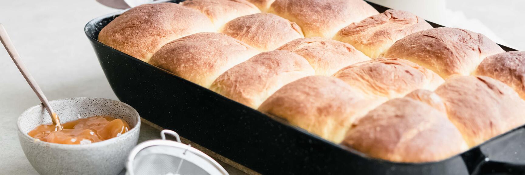 Baked buns in black baking tray with jam bowl and powdered sugar sieve beside.