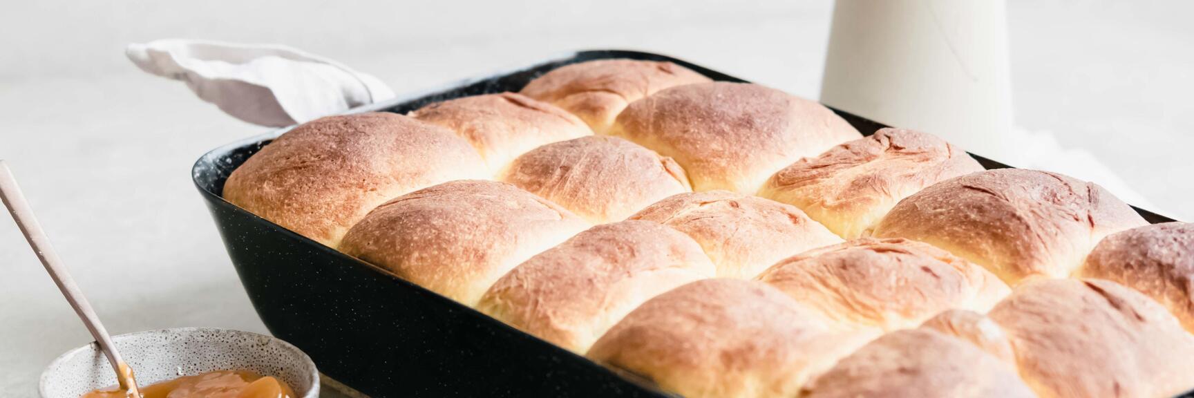 Buchteln in schwarzem Backblech, daneben Schüssel mit Marmelade und Puderzucker-Sieb.