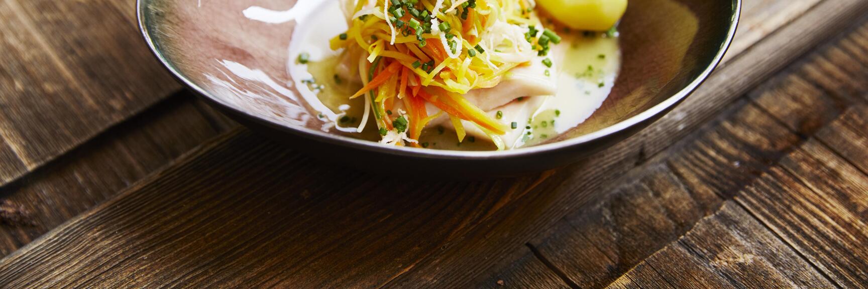 Close-up: meat dish in dark bowl with vegetable strips, chives, cheese and potato on wooden table.