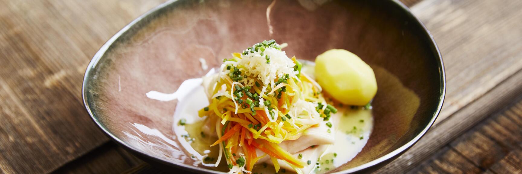 Nahaufnahme: Fleischgericht in dunkler Schale mit Gemüsestreifen, Schnittlauch, Käse und Kartoffel auf Holztisch.