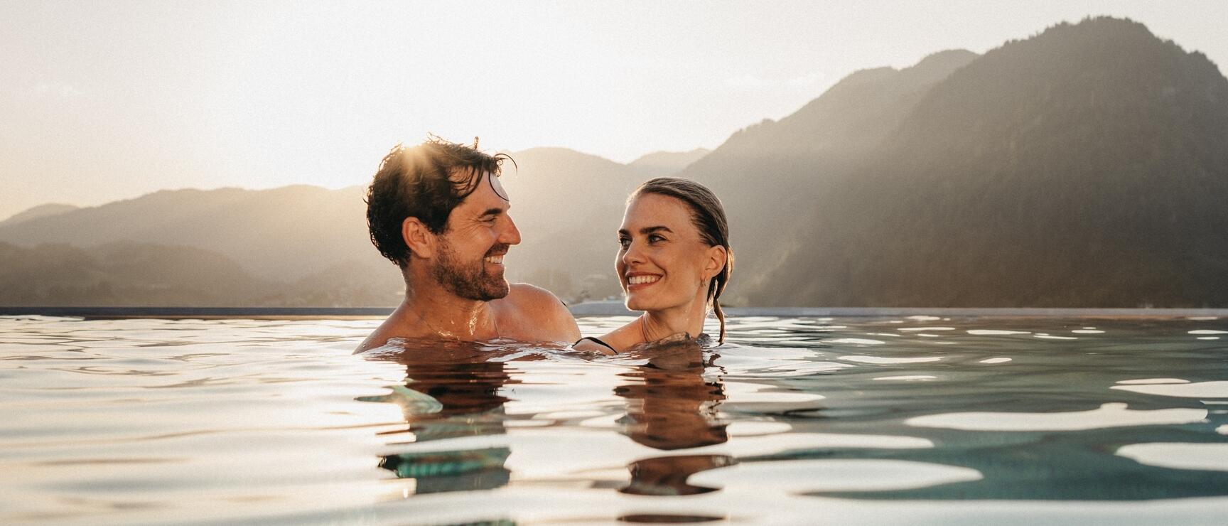 Two laughing people in an infinity pool with mountain panorama backlit.