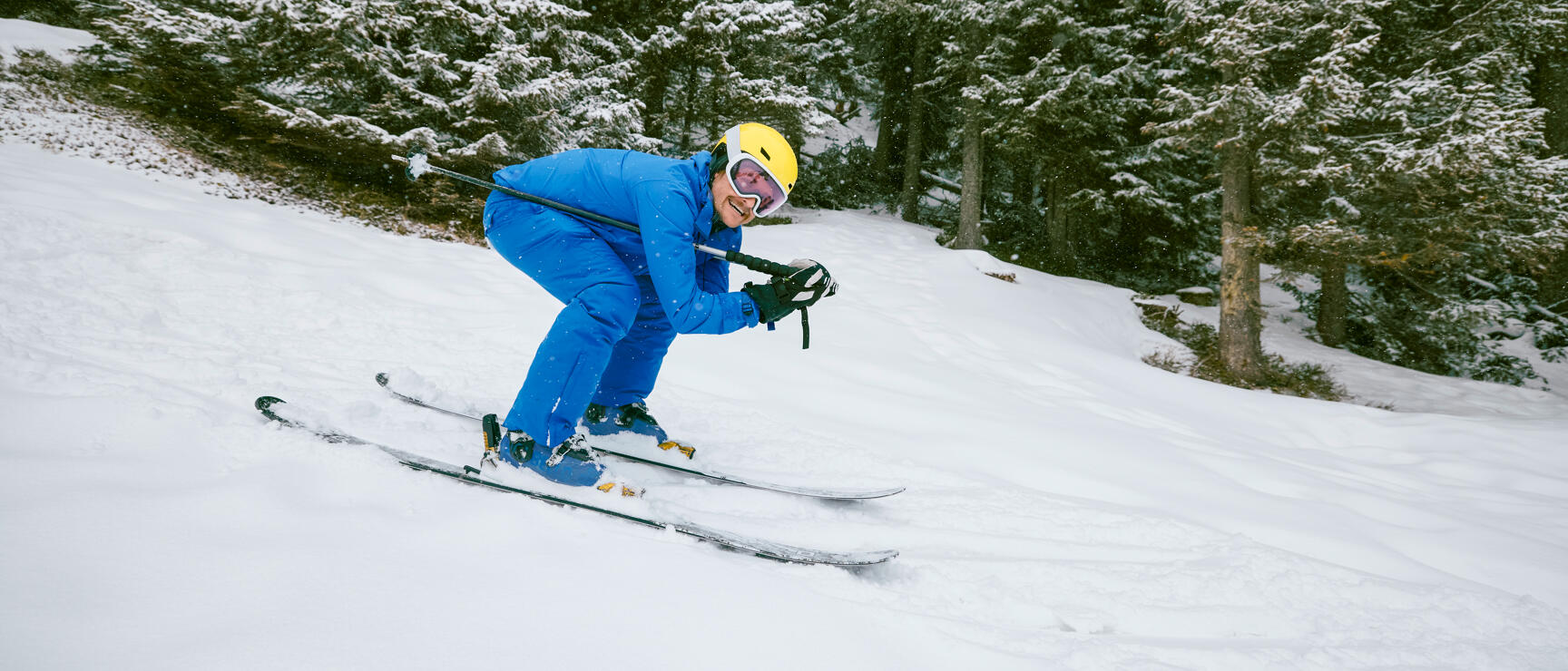 Skifahrer in blauem Anzug und gelbem Helm fährt Steilhang hinunter, verschneite Tannen im Hintergrund.
