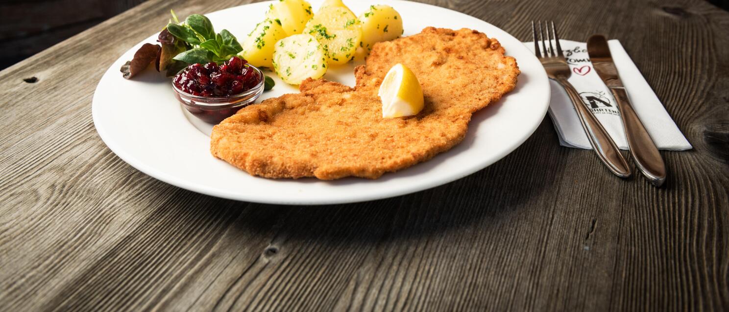 Wiener Schnitzel with lemon, parsley potatoes and lingonberries on white plate, wooden table.