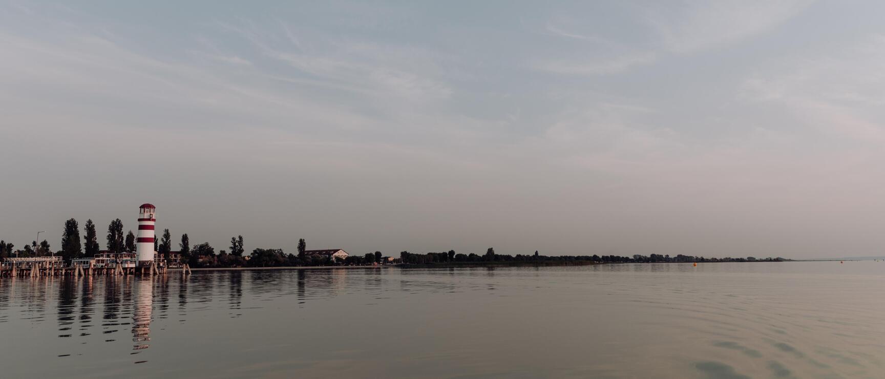 White lighthouse with red stripes at Lake Neusiedl harbour, calm water, flat shoreline.