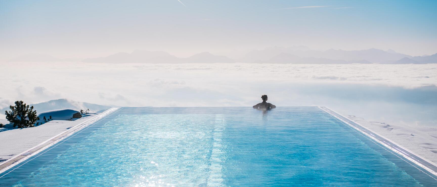 Person in infinity pool above sea of clouds, snow-covered mountains in background, blue sky.