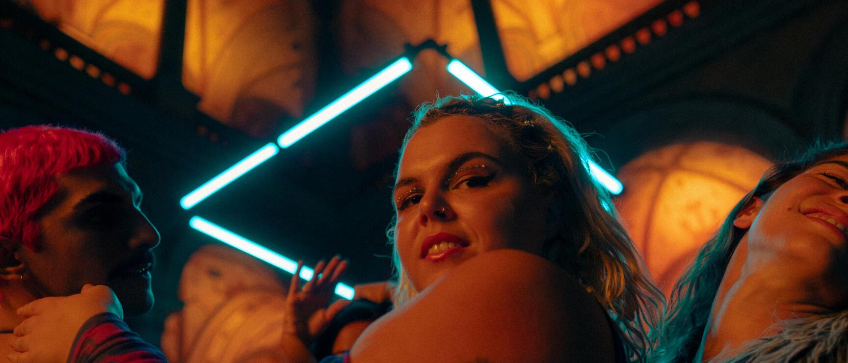 Tanzende Personen in einem Club mit türkisfarbenem Neonlicht, Glitzer-Make-up und Tätowierungen.