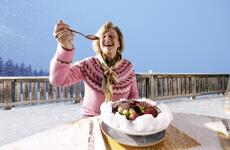 Smiling woman with spoon in front of a bowl on a snowy alpine terrace, blue winter sky.