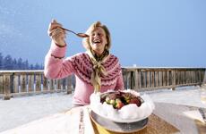 Smiling woman with spoon in front of a bowl on a snowy alpine terrace, blue winter sky.