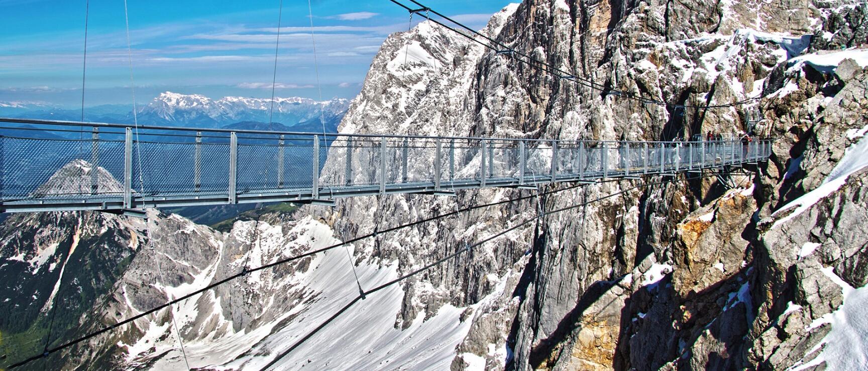 Steel suspension bridge between snow-capped rock peaks at Dachstein, alpine panorama in background.