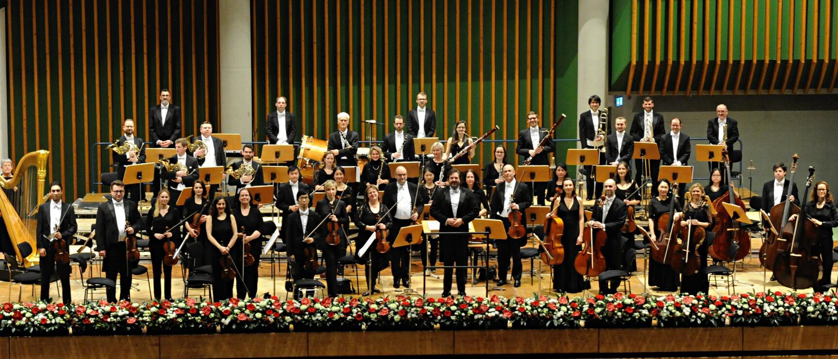 Large orchestra in black concert attire on stage with music stands and red flowers.