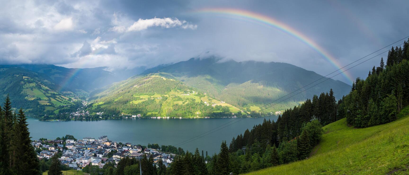 Panoramic view of Zell am See with Zeller See lake, green mountains and rainbow in cloudy sky.