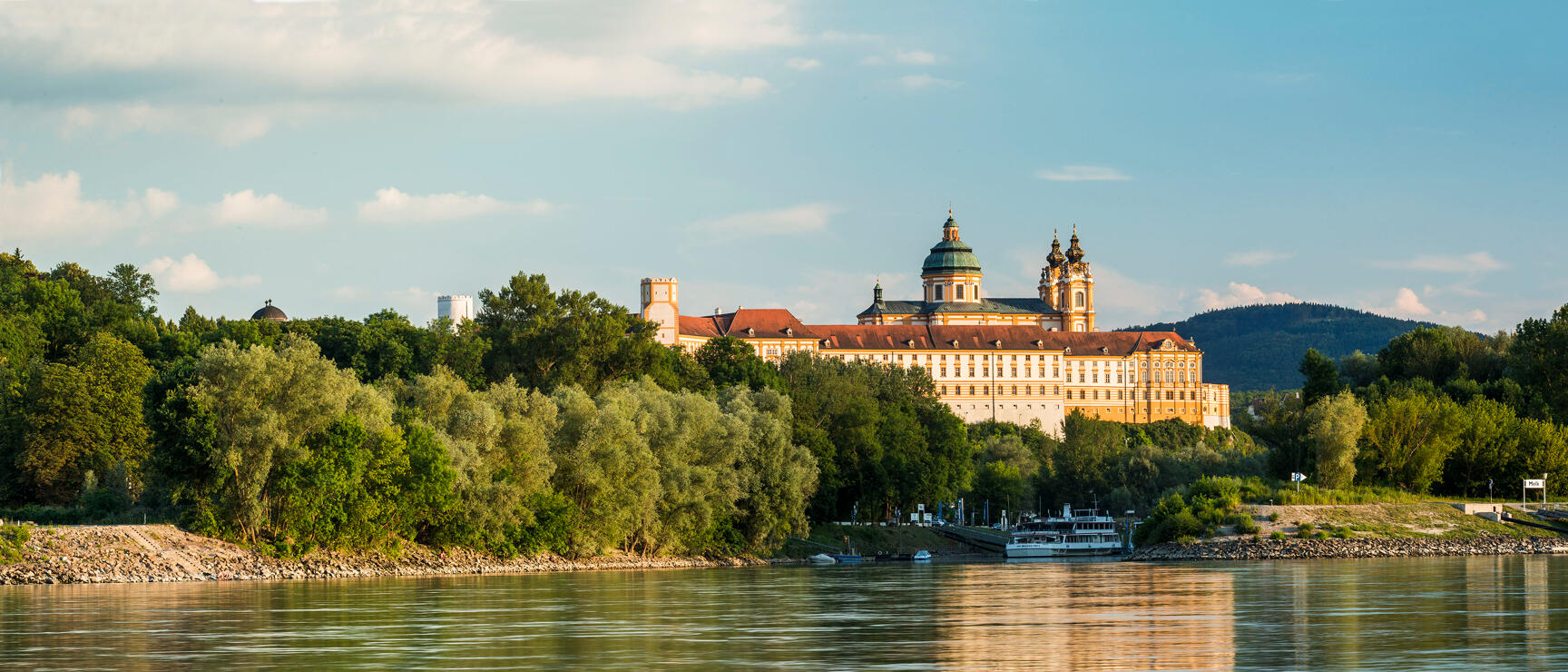 Melk Abbey perched on a rock above the Danube, ship at the shore, trees in foreground, blue sky.