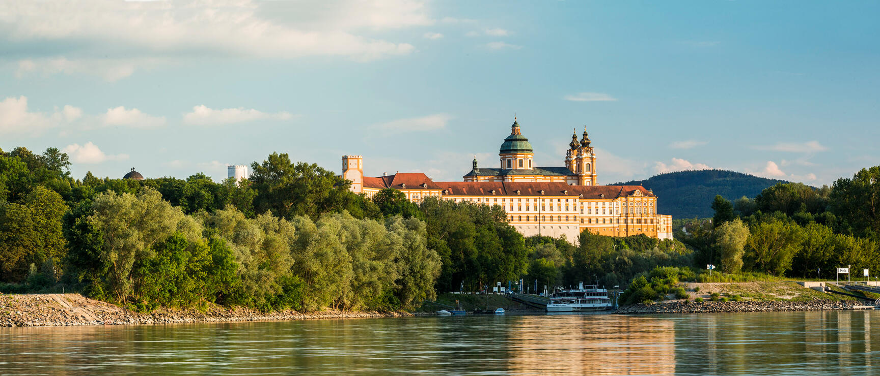 Melk Abbey perched on a rock above the Danube, ship at the shore, trees in foreground, blue sky.
