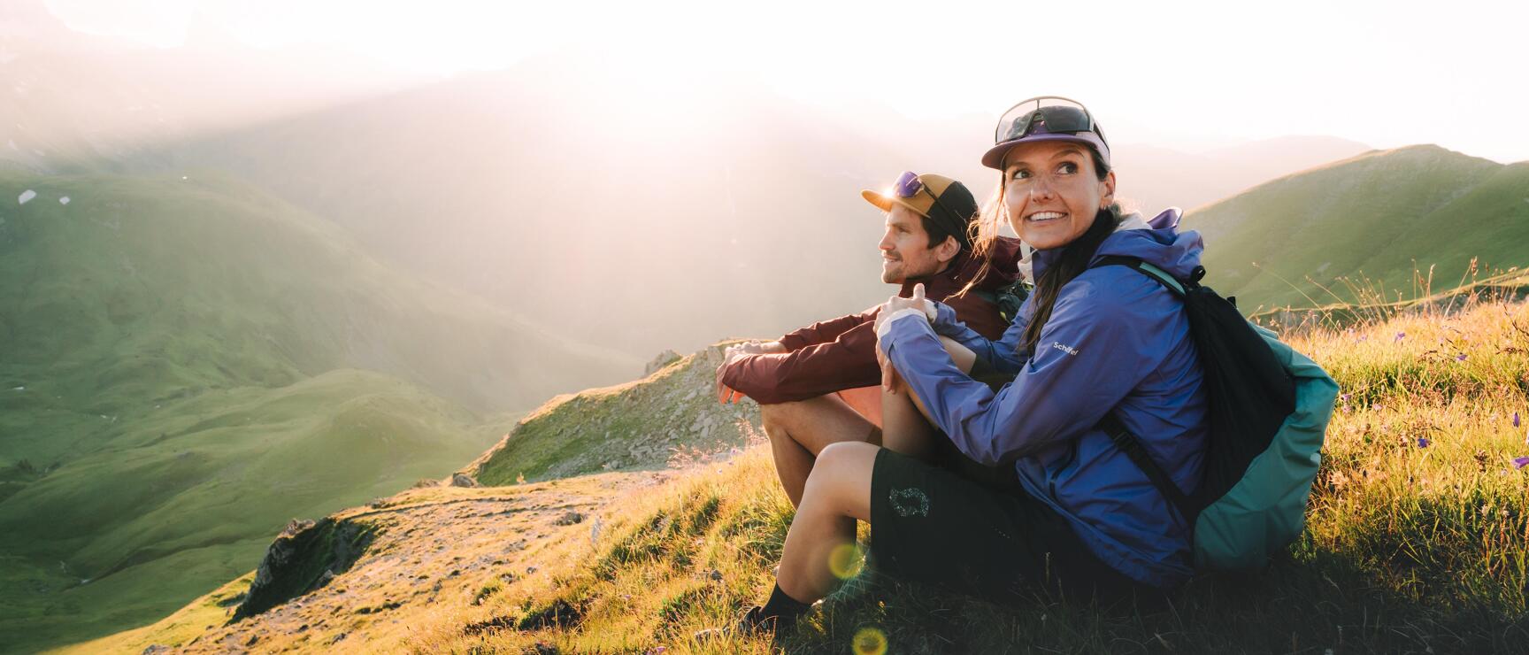 Two hikers wearing caps sit on a grassy slope, green mountain peaks backlit in the background.