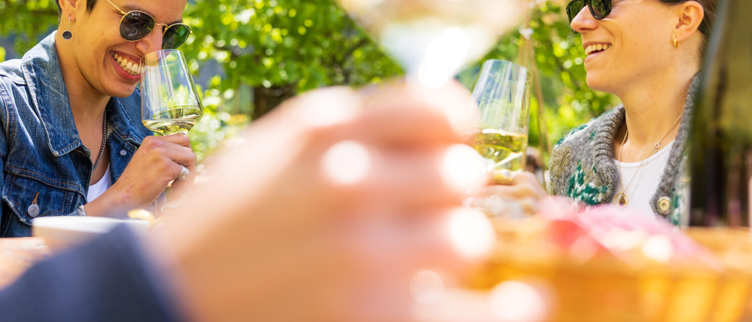 Zwei lächelnde Frauen mit Sonnenbrille stoßen im Freien mit Weißwein an, grüne Bäume im Hintergrund.