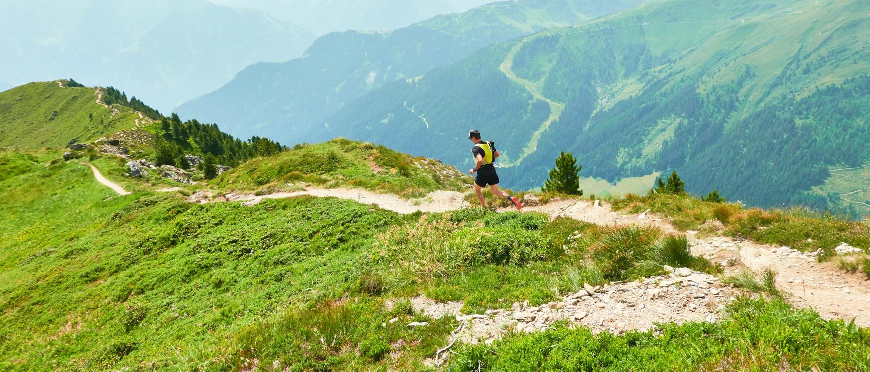 Trail runner with backpack on a mountain path, wide alpine panorama with green slopes in the background.