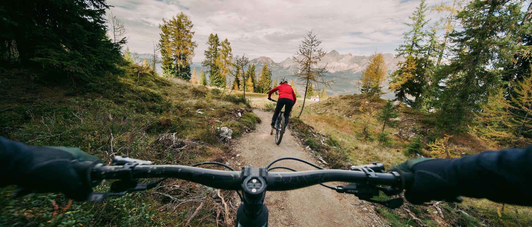 POV shot: mountain bike handlebars, person ahead in red jacket on gravel path, mountain panorama and autumn larches.
