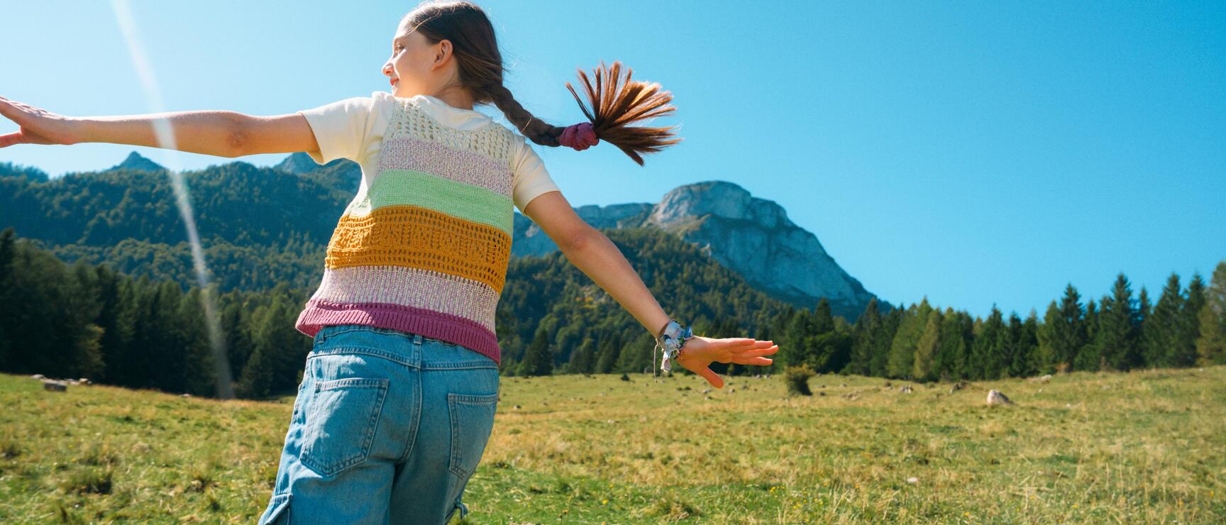 Girl with outstretched arms on alpine meadow with mountain landscape and blue sky in background.