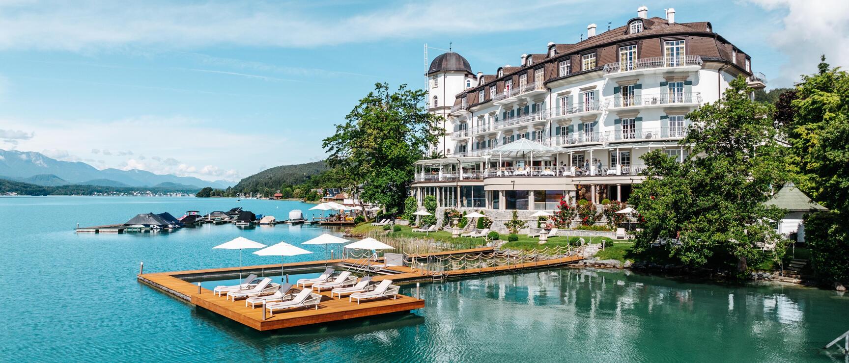 White grand hotel at Lake Wörthersee with wooden dock, loungers and sun umbrellas, mountains in background.