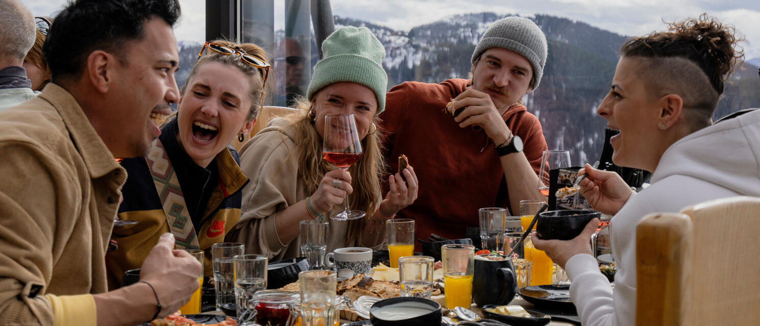 Five people dining at a wooden table, wine glasses, orange juice, snow-capped mountains through panoramic windows.